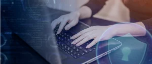 Person typing on a laptop keyboard with blue code graphics and a shield symbol, conveying cybersecurity.