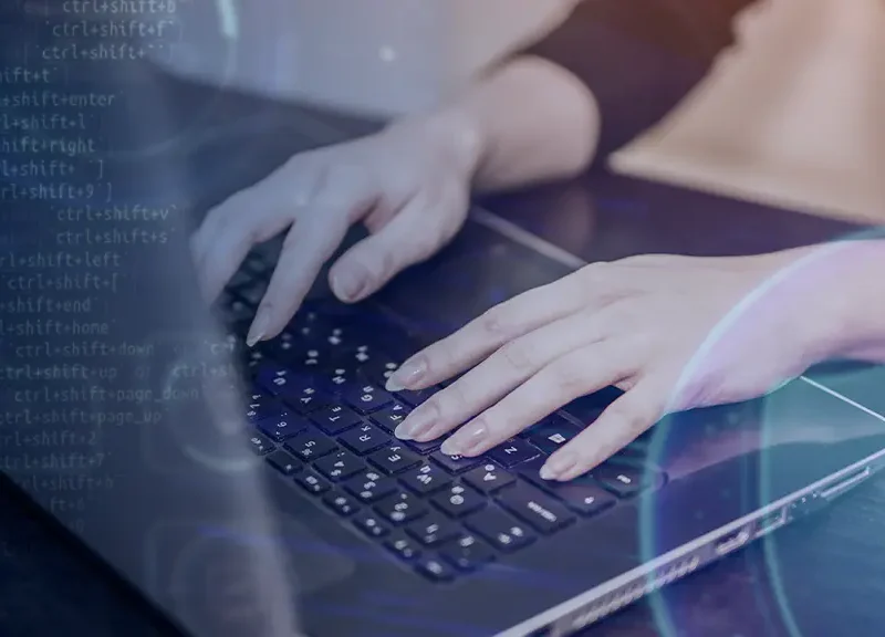 Person typing on a laptop keyboard with blue code graphics and a shield symbol, conveying cybersecurity.